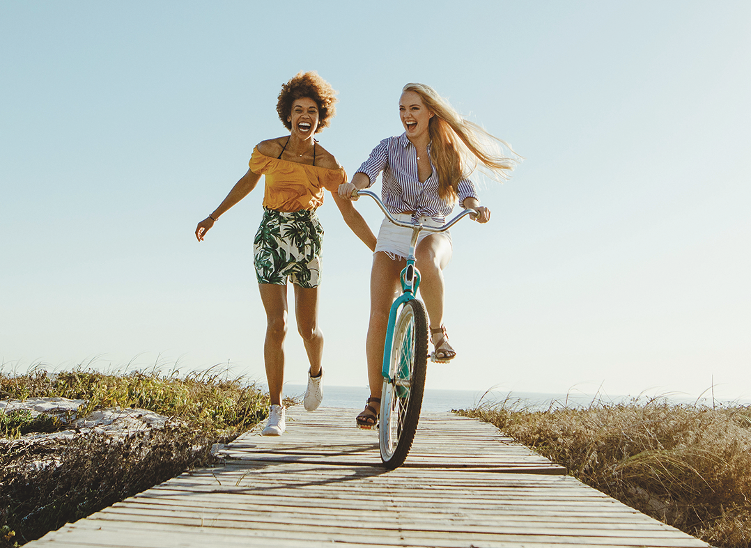 Eine Frau rennt, eine andere Frau sitzt auf einem Fahrrad. Beide lachen.